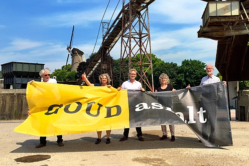 Foto: Op het industrieterrein van de voormalige Koudasfaltfabriek houden Peterpaul Kloosterman samen met andere betrokken inwoners een vlag op met een goudkleurig en een zwart vlak met daarop de tekst GOUDasfalt